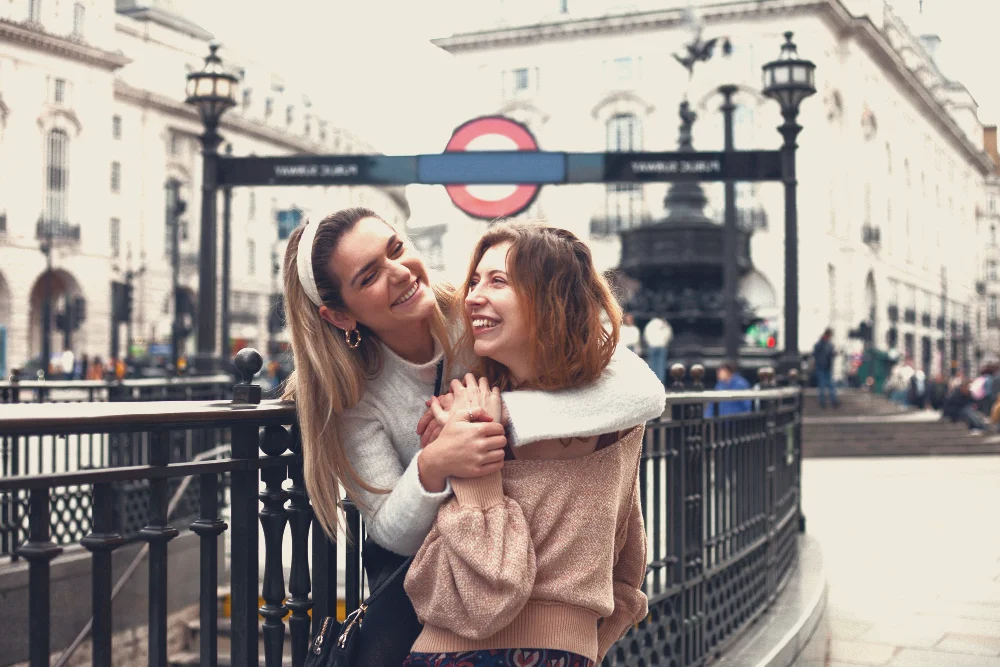 Two women laughing and hugging in front of a tube entrance after they got their indefinite leave to remain visa