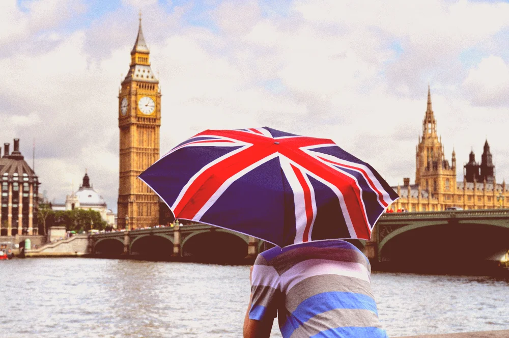 A tourist man holding an umbrella with the United Kingdom flag on it, looking at the Big Ben near the River Thames.