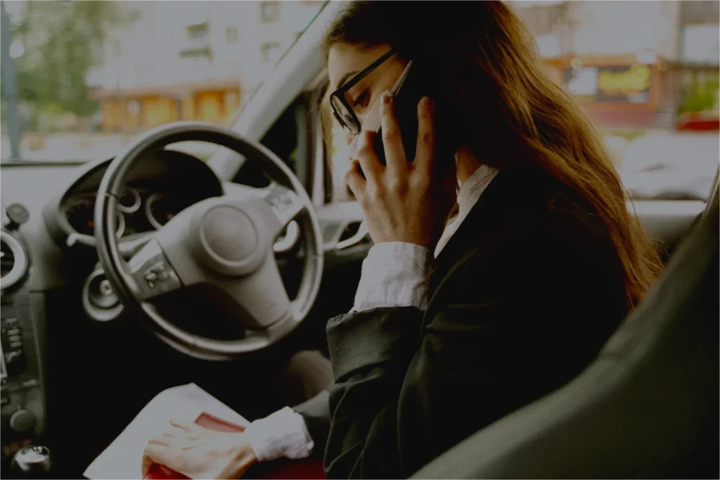 A woman sitting in the driver's seat, talking on the phone and holding a document despite having no license and no insurance.